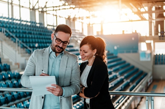 Dois profissionais a sorrirem enquanto analisam apontamentos num bloco de notas, numa bancada desportiva vazia, em contexto de planeamento e organização de um evento.