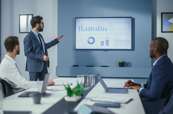 Profissional em fato a apresentar gráficos e dados de desempenho numa reunião de negócios, com colegas sentados à mesa a observar a apresentação num ecrã de parede.